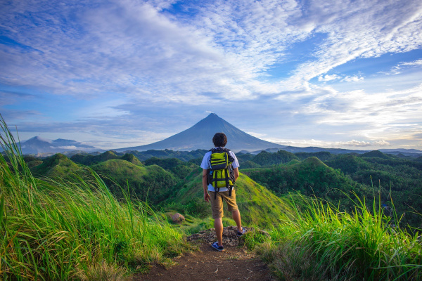 man standing in lush green fields looking ahead at a volcano, framed by blue skeis