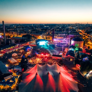 Tents and stages within an apparent former industrial park are illuminated by colorful lights and overflowing with visitors. In the background the sun has just set on the city skyline.